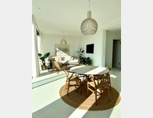 Helles Wohnzimmer mit modernen Holz- und Naturakzenten; ein runder Esstisch mit Holzstühlen steht auf einem geflochtenen Teppich, daneben eine Sitzecke mit Sofa und Pflanzen. Zwei hängende Lampen und ein Fernseher an der Wand ergänzen die minimalistische Einrichtung.