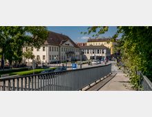 Städtische Szene mit einem historischen Gebäude mit hellem Putz und Walmdach im Hintergrund, flankiert von moderneren Gebäuden. Im Vordergrund ist eine eingerahmte Gehwegbrücke zu sehen, die von Bäumen und Grünflächen umgeben ist.