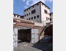 Gebäudeensemble mit einem weißen, mehrstöckigen Gebäude im Hintergrund und einer Backsteinmauer mit Metalltoren und Außentreppe im Vordergrund. Der gepflasterte Hof wird von Ziegelarchitektur und einem klaren Himmel umgeben.