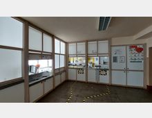 Essensausgabe in einer Kantine mit einer Trennwand aus Milchglas und Holzrahmen. Dahinter sind zwei Essensausgaben mit Schildern ("Essen 1" und "Essen 2"), sowie eine Küche mit gelben Fliesen sichtbar. Auf dem Boden markieren gelbe Klebebänder Laufwege.