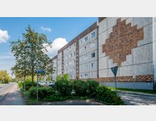 Mehrstöckiges Wohnhaus aus Betonplatten mit geometrischem Mosaikmuster aus Ziegelsteinen an der Seitenfassade. Im Vordergrund befinden sich ein Gehweg, ein Baum, Straßenschilder und kleine Grünflächen.