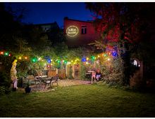 Ein beleuchteter Garten in der Nacht mit bunten Lichterketten, diese hängen zwischen Bäumen und umgeben einen Sitzbereich mit Metallstühlen und -tischen. Im Hintergrund ist eine Fassade mit einem leuchtenden Schild „Ballhaus Wedding“ und dichter Vegetation sichtbar.