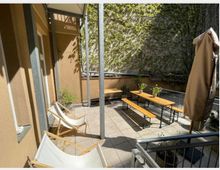 Sonniger Innenhof mit gepflastertem Boden, Holzbänken und Tischen, sowie zwei weißen Liegestühlen. Eine mit Efeu bewachsene Wand bildet den Hintergrund, während eine beige Hausfassade und ein verschlossener Sonnenschirm zu sehen sind.