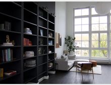 Wohnraum mit einer hohen Fensterfront, die viel Tageslicht hereinlässt. Links stehen schwarze Bücherregale, rechts befindet sich ein Sitzbereich mit einer weißen Couch, einem runden Holztisch und einem Sessel; eine Zimmerpflanze und minimalistische Dekoration komplettieren den Raum.