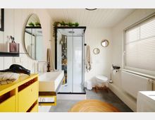 Helles Badezimmer mit weiß vertäfelten Wänden, einer Glasdusche mit schwarzem Rahmen, einem weißen Waschbecken und einer gelben Kommode. Ein großes Fenster mit Jalousien, kreisförmige Wandspiegel und ein runder, orangefarbener Teppich ergänzen die Einrichtung.