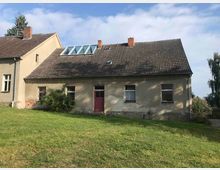 Ein einstöckiges Wohnhaus mit beigem Putz und einem dunklen Satteldach, teils mit Dachfenstern. Im Vordergrund befindet sich eine grüne Rasenfläche, umgeben von vereinzelten Sträuchern und Bäumen.