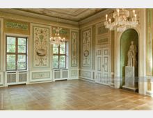 Eleganter historischer Raum mit verziertem Parkettboden, Wänden mit klassischen Wandmalereien in Grün- und Beigetönen, Kronleuchtern und großen Fenstern mit Blick ins Grüne. Rechts steht eine Skulptur in einer Wandnische, die von dekorativen Ornamenten umgeben ist.