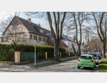 Straßenszene in einem Wohngebiet mit gepflastertem Gehweg und parkenden Autos am Straßenrand. Auf der linken Seite sind mehrere zweistöckige Häuser mit Satteldächern, umgeben von Bäumen und Hecken. Straßenschilder kennzeichnen die Kreuzung von Drygalski- und Schweinfurthstraße.