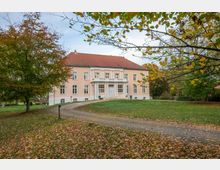 Herrenhaus mit rosa Fassade und rotem Ziegeldach, umgeben von einem herbstlichen Park mit Laubbäumen und einem geschwungenen Kiesweg, der zum Haupteingang mit Säulenportikus führt.