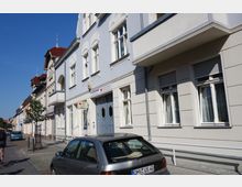 Straßenszene mit historischen Mehrfamilienhäusern, teils mit Zierelementen, und einem Gebäude, das ein Schild des Deutschen Roten Kreuzes mit der Aufschrift „Pflegedienst“ trägt. Im Vordergrund parken Autos am Straßenrand, während eine Radfahrerin auf dem Gehweg zu sehen ist.