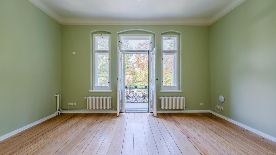 Leerer Wohnraum mit hellgrünen Wänden, hohen Holz-Dielenböden und drei hohen Fenstern, in der Mitte eine geöffnete Balkontür mit schmiedeeisernem Geländer. Unter den seitlichen Fenstern stehen Heizkörper; die Decke ist weiß mit einfachem Deckenanschluss und zentraler Leuchte.