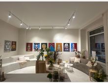 Heller, minimalistischer Galerieraum mit weißer Sitzstufe und grauem Sofa, beleuchtet durch Deckenschienen-Spots. An der Rückwand hängt eine Reihe farbiger Gemälde, davor stehen Baumstamm-Tische, kleine Beistelltische und mehrere Vasen mit Blumen; rechts ein großes Fenster zur Straße.