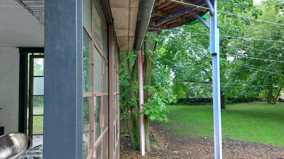 Außenansicht mit einer verglasten Fassade und einem grauen Metallrahmen, daneben ein Überdach mit sichtbarer Metallstütze. Im Hintergrund ist eine Grünfläche mit Bäumen und Sträuchern zu sehen.