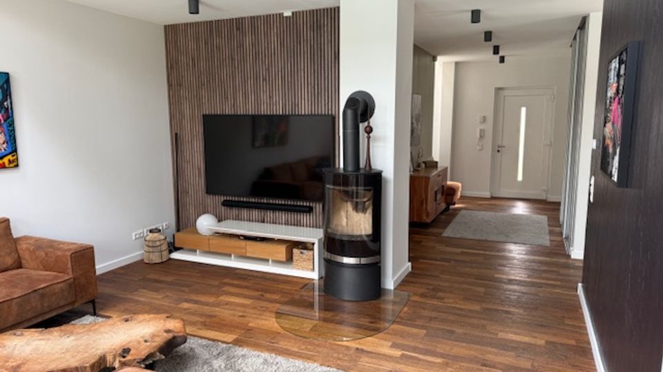 Modern open-plan living room and hallway with hardwood floors, a wall-mounted TV on a vertical wood-slat accent wall, and a freestanding cylindrical black stove on a glass hearth. Recessed ceiling lights line the corridor leading to a white front door with a narrow vertical window.