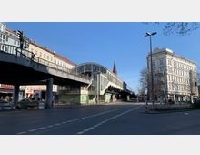 Straßenszene mit einer erhöhten U-Bahn-Station im Zentrum, die durch große Fenster und ein geschwungenes Dach gekennzeichnet ist. Rechts stehen historische Wohngebäude mit ornamentaler Fassade, während links Geschäfte und geparkte Autos zu sehen sind; im Hintergrund ragt eine Kirchturmspitze hervor.