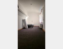 Schmaler Raum mit weiß gestrichenen Wänden, einem dunklen Teppichboden und einem Einzelbett auf einem Metallgestell an der hinteren Wand. Rechts befindet sich ein Fenster mit durchscheinendem Vorhang, darunter steht ein Heizkörper.