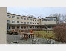 Innenhof einer modernen, hell verputzten Gebäudeanlage mit drei Stockwerken und vielen rechteckigen Fenstern, rechts mit einem abgerundeten Gebäudeteil. Im Vordergrund stehen Holz-Picknicktische auf gepflasterter Terrasse neben einer Rasenfläche mit Sträuchern, Pflanzkübeln und einzelnen Bäumen.