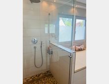 Moderne Badezimmer-Dusche mit weißen Fliesen, einer bodengleichen Duschkabine aus Glas und einem mosaikartigen Bodenbelag. Im Hintergrund befindet sich ein Fenster mit Milchglas und eine gepolsterte Liegefläche mit Kissen.