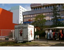 Ein kleiner Rostbratwurst-Imbisswagen steht auf einer Grünfläche vor mehrstöckigen Gebäuden, darunter ein orangefarbenes und ein graues Gebäude mit großen Fenstern. Rechts neben dem Imbisswagen befindet sich ein Verkaufsstand mit Kleidung unter einem weißen Pavillondach.