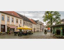Historischer Marktplatz mit Kopfsteinpflaster, umgeben von Häusern mit pastellfarbenen Fassaden. Im Vordergrund das "Lindower Stadtcafé" mit gelben Sonnenschirmen und Sitzgelegenheiten sowie ein großer Baum und ein Blumenstand.