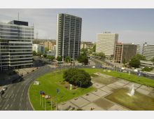 Stadtplatz mit einer großen Rasenfläche, einem zentralen Baum, mehreren Flaggenmasten und Wasserspielen im Vordergrund. Umgeben von mehrspurigen Straßen und modernen Bürogebäuden, darunter ein Hochhaus der Technischen Universität.