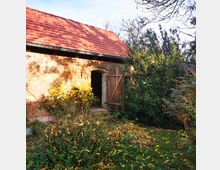 Backsteingebäude mit rotem Ziegeldach, einer geöffneten Holztür und einem überwucherten Garten mit herbstlichem Laub im Vordergrund. Dichte Sträucher und Bäume umgeben das Gebäude.