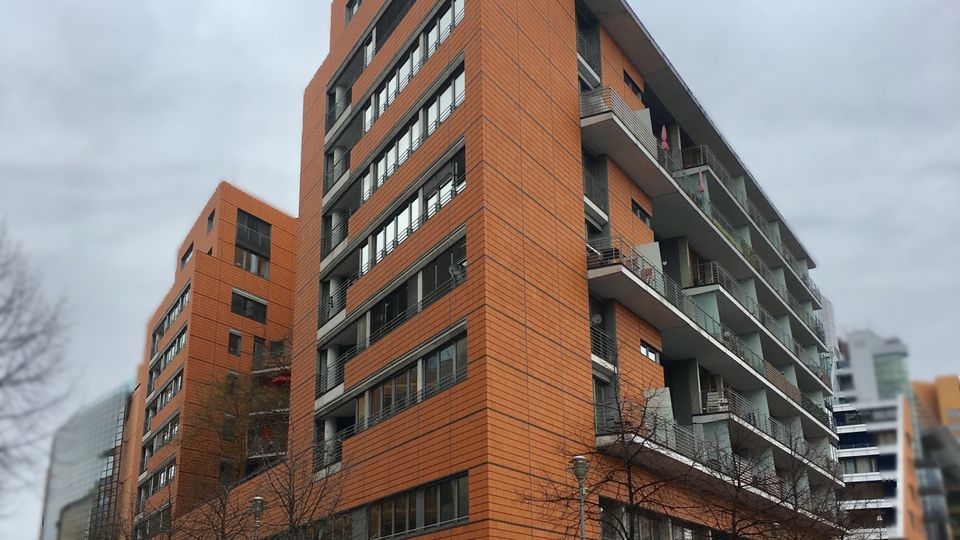 Moderner, mehrstöckiger Wohn- und Bürokomplex mit orangefarbener Fassadenverkleidung, großen Fensterbändern und durchgehenden Balkonen an der Straßenecke. Im Vordergrund verlaufen breite Gehwege mit kahlen Straßenbäumen, Fahrradständern und Straßenbeschilderung an einer Kreuzung.