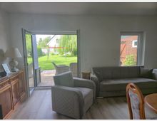 Helles Wohnzimmer mit grauem Sofa und Sessel sowie Holzkommode; eine große, geöffnete Terrassentür führt auf eine gepflasterte Terrasse und in einen grünen Garten. Draußen sind Rattanmöbel, ein Rasen mit Schaukelgestell und Bäume zu sehen.