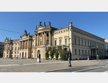 Historisches Gebäude mit klassizistischer Architektur, vertreten durch Säulen, dekorative Statuen auf dem Dach und zahlreiche Fenster. Das Gebäude steht an einem gepflasterten Platz bei klarem Himmel, flankiert von Laternenpfählen und Passanten.
