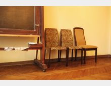 Ein schlicht eingerichteter Raum mit einem hölzernen, mobilen Schultafelständer auf der linken Seite und drei Stühlen mit gemusterten beziehungsweise einfarbigen Polstern an einer gelb gestrichenen Wand. Der Boden besteht aus Holzparkett.