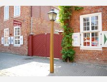 Backsteingebäude mit weißen Sprossenfenstern und einem dunkelroten Holztor, daneben eine Holzlaterne und Kletterpflanzen an der Wand; Pflastersteine bedecken den Boden.