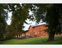 Backsteinbau mit mehreren rechteckigen Fenstern und einer Terrasse, die mit Tischen und geschlossenen Sonnenschirmen ausgestattet ist. Das Gebäude liegt in einer grünen Parklandschaft, umgeben von großen Bäumen.