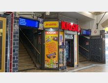 Ein kleiner Kiosk in einem Bahnhof mit rotem Leuchtschild „KIOSK“. Neben der Eingangstür befindet sich ein Plakat mit Werbung für Snacks, Getränke und Tabakwaren. Im Hintergrund sind Treppen, eine Bahnhofsanzeige und Wandfliesen in dunkelblau zu sehen.