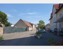 Straßenszene in einem ruhigen Dorf mit mehreren Wohnhäusern mit roten Ziegeldächern und heller Fassade. Ein Teil der Straße ist durch Bauzäune abgesperrt, ein geparktes Auto und Pflanzkübel stehen im Vordergrund.