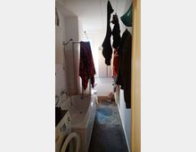 Ein schmaler Badezimmerraum mit einer Badewanne auf der linken Seite und einer Waschmaschine im Vordergrund. Über der Badewanne und dem Gang hängen Kleidung und Handtücher zum Trocknen auf Wäscheleinen. Im Hintergrund ist ein Fenster zu sehen, das Tageslicht hereinlässt.