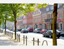 Straßenansicht mit historischen Backsteingebäuden und kleinen Geschäften, parkenden Autos und Bäumen, die entlang eines Gehwegs Schatten spenden. Im Hintergrund ist leichte städtische Bebauung zu sehen.