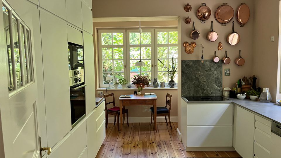 Helle Wohnküche mit weißen Einbauschränken, Holzfußboden und einer U-förmigen Arbeitsfläche mit Marmor-Rückwand. Vor großen Sprossenfenstern steht ein kleiner Holztisch mit zwei Stühlen, an der Wand hängen Kupfertöpfe und -pfannen.