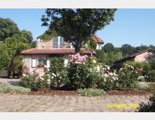 Ein kleines Backsteinhaus mit roten Dachziegeln, von rankendem Grün bewachsen, steht inmitten eines Gartens mit blühenden Rosensträuchern. Im Vordergrund ist ein gepflasterter Weg zu sehen, umgeben von Bäumen und weiteren Pflanzen.