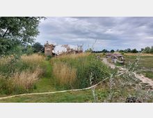 Eine ländliche Szene mit einer aus Holz und Planen improvisierten Hütte, umgeben von hohem Gras und Gebüsch. Ein unbefestigter Weg aus Holzplanken führt durch die Landschaft, die von Wiesen und vereinzelten Bäumen geprägt ist, unter einem bewölkten Himmel.