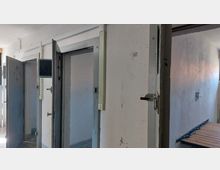 Flur in einem Gebäude mit geöffneten Metalltüren, die verstärkt wirken und mit Schlössern sowie kleinen Sichtfenstern versehen sind; rechts im Bild befindet sich ein einfaches Bettgestell aus Holz und Metall.