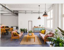Wohnzimmer im skandinavischen Stil mit hellen Wänden und großen Fenstern, die viel Tageslicht hereinlassen. Zu sehen sind zwei rosa Sofas mit bunten Kissen, ein hölzerner Couchtisch auf einem beigen Teppich, viele Zimmerpflanzen und eine türkisfarbene Kommode; im Hintergrund befindet sich ein Esstisch mit Holzstühlen.