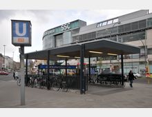 Außenansicht der U-Bahn-Station "Walther-Schreiber-Platz" in Berlin, mit überdachtem Eingangsbereich, Fahrradständern und umliegenden Geschäften, darunter ein modernes Einkaufszentrum mit Glasfassade. Im Vordergrund steht ein Schild mit dem Logo der U-Bahn-Linie U9.