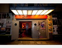 Eingang eines Clubs oder Veranstaltungsortes mit einem rot beleuchteten Vordach und der Aufschrift "Lido". Die grauen Türen mit Bullaugen sind geöffnet und geben den Blick auf einen beleuchteten Innenraum frei; die Außenwände sind mit Graffiti und Plakaten bedeckt.