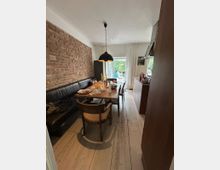 Esszimmer mit Holzboden und einer Backsteinwand auf der linken Seite. Ein Holztisch mit Stühlen steht im Zentrum, darüber hängt eine schwarze Hängeleuchte; im Hintergrund ist eine Glastür zu einem Garten sichtbar.