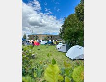 Eine Wiese mit mehreren aufgebauten Zelten, umgeben von Büschen und Bäumen. Im Hintergrund steht ein modernes, halbkreisförmiges Gebäude mit großen Fenstern unter einem bewölkten Himmel.