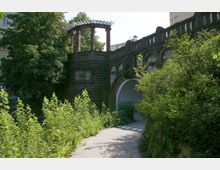 Historische Steinstruktur mit einem runden Pavillon mit Säulen und einem gewölbten Durchgang, umgeben von dichter Vegetation und hohen Bäumen. Im Hintergrund sind moderne Gebäude zu sehen.