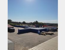 Freigelände eines Industrie- oder Lagerbereichs mit mehreren blauen Schiffscontainern, einer gepflasterten Fläche und gelagerten zylindrischen Gegenständen. Im Hintergrund sind Bäume und eine ländliche Landschaft unter klarem Himmel erkennbar.