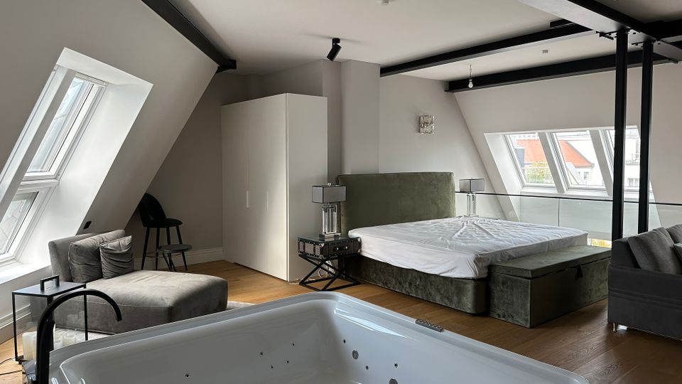 Modernes Dachgeschoss-Schlafzimmer mit schrägen Wänden, sichtbaren schwarzen Stahlträgern und großen Dachfenstern. Im Raum stehen ein Bett mit gepolstertem Kopfteil, ein Kleiderschrank, Sitzmöbel und im Vordergrund eine eingelassene Whirlpool-Badewanne auf Holzboden.