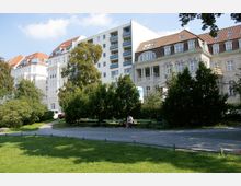 Ansicht eines kleinen Parks mit grünen Rasenflächen, Bäumen und Gehwegen, umgeben von historischen und modernen Wohngebäuden. Im Vordergrund stehen grüne Parkbänke, von denen eine von einer Person genutzt wird.