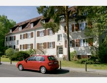 Zweistöckiges Wohngebäude mit weißer Fassade, braunen Fensterläden und einem roten Satteldach. Im Vordergrund steht ein rotes Auto auf der Straße, umgeben von Bäumen und gepflegtem Grün.